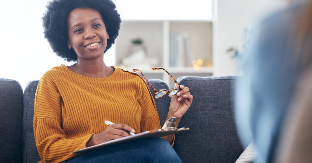 A smiling therapist holds her glasses and a clipboard while listening to a client during a mental health services session.