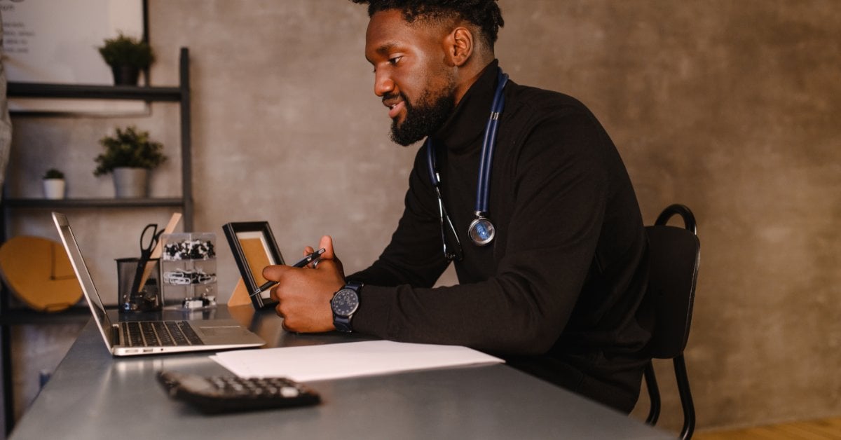 A male mental health professional sits at his office desk with a laptop, engaged in a virtual therapy session with a patient.