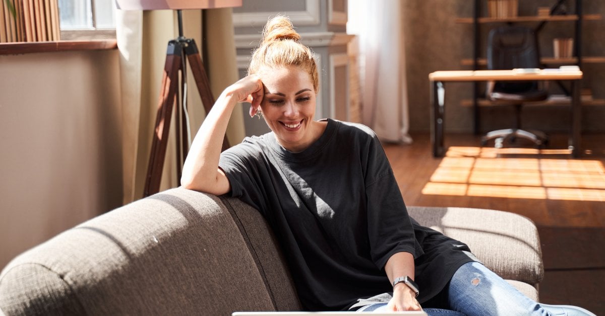 A smiling woman sits on her couch with a laptop, engaged in an online therapy session from the comfort of her home.