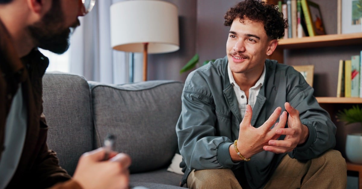 A smiling young man gestures as he speaks with a mental health professional during a therapy session in a cozy office.