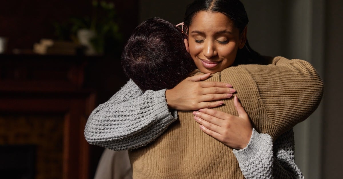 Two women share a warm embrace indoors, one smiling with her eyes closed, capturing the comfort of human connection.