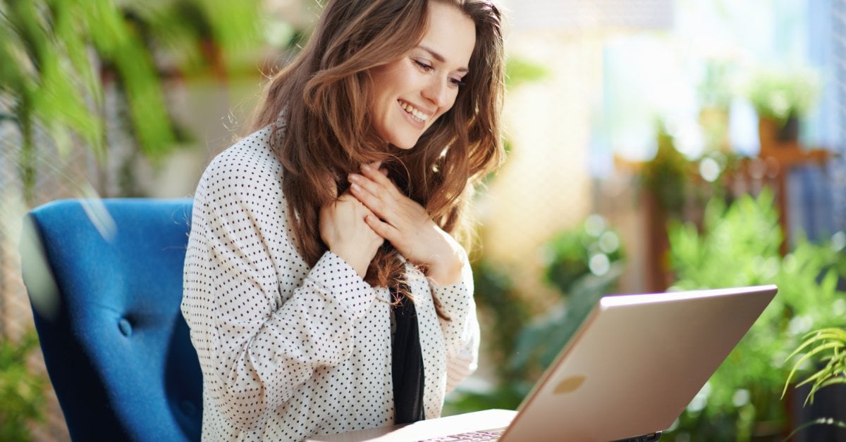 A smiling woman sits in a sunlit room with plants, looking at her laptop during an online telehealth therapy session.