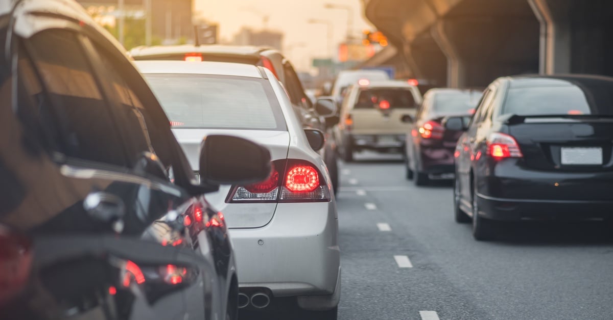 Rows of cars with glowing brake lights sit in slow-moving highway traffic beneath an overpass at sunset.