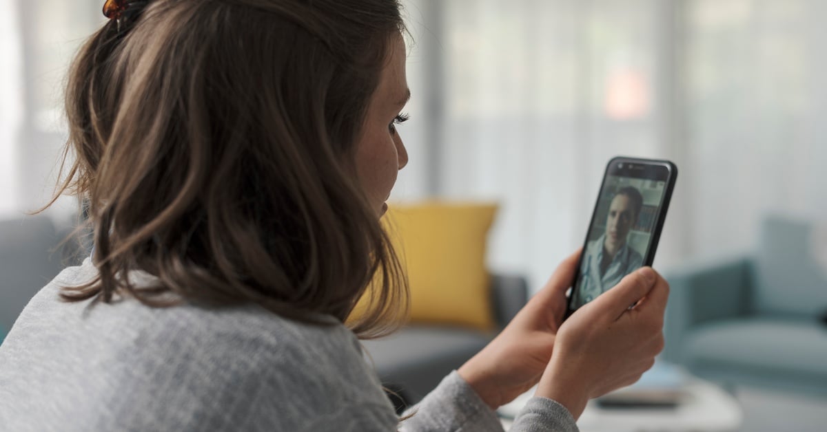 A woman holds her smartphone and attends a telehealth video appointment with a doctor from her living room.