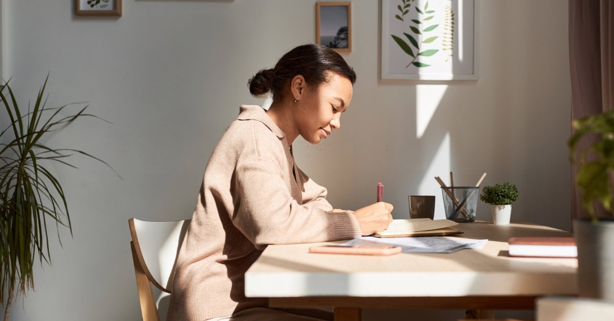 A woman journals at a sunlit table between therapy sessions, writing in a notebook surrounded by plants and books
