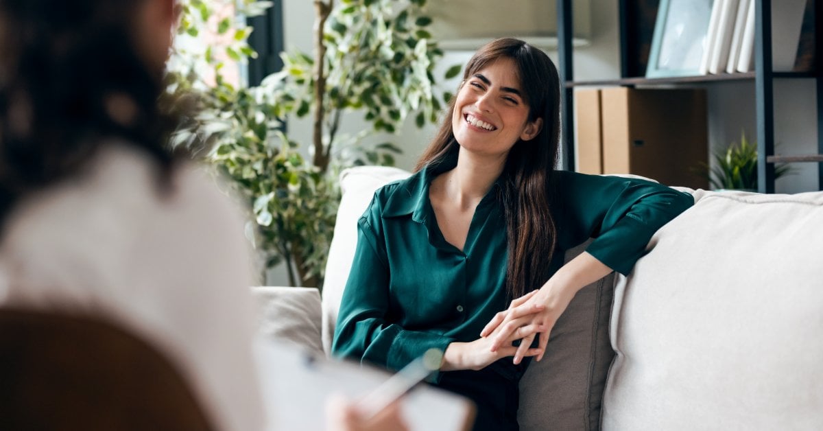 A smiling young woman talks with her therapist, who takes notes, during a mental health counseling session.