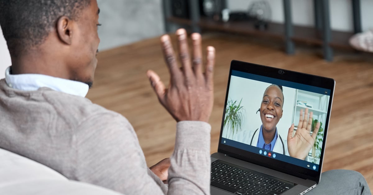 A man sitting at home waves to a smiling doctor on a laptop screen during a telehealth video appointment.