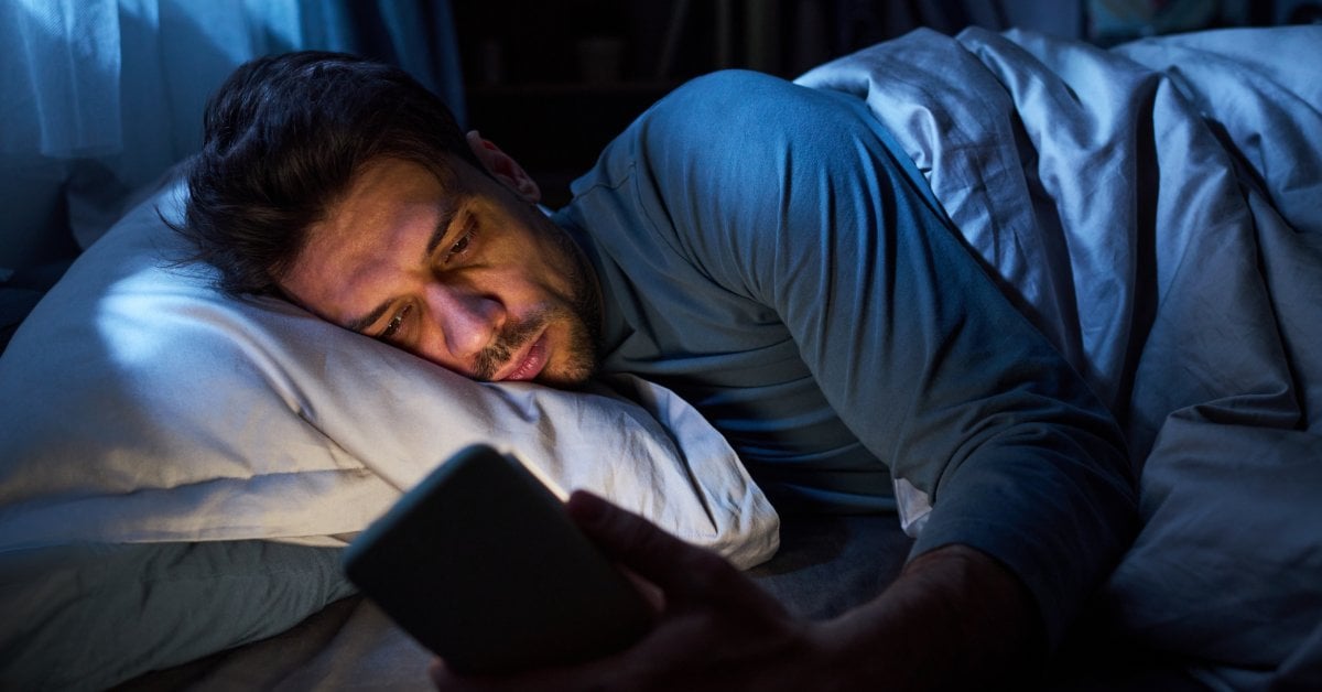 At night, a man lies on his side in bed while looking at a smartphone. The screen's light illuminates his face.