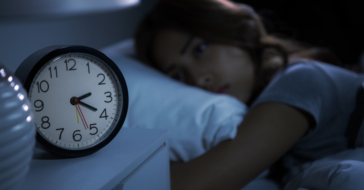 An analog clock on a bedside table shows 2:20. A blurred woman lies on her side in bed, her eyes open in the dark.