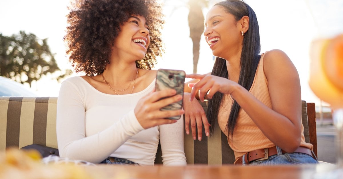 Two women sit together outdoors, smiling and laughing. One holds a smartphone while the other gestures toward it.