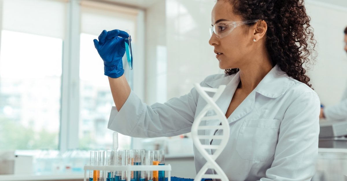 A lab worker holds up a filled test tube to look at it. More test tubes and a DNA-shaped model are on the lab bench.