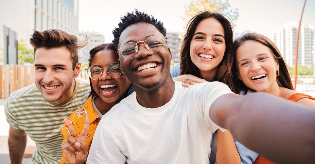 Five smiling young adults pose closely together for a selfie outdoors. Tall buildings and landscaping are behind them.