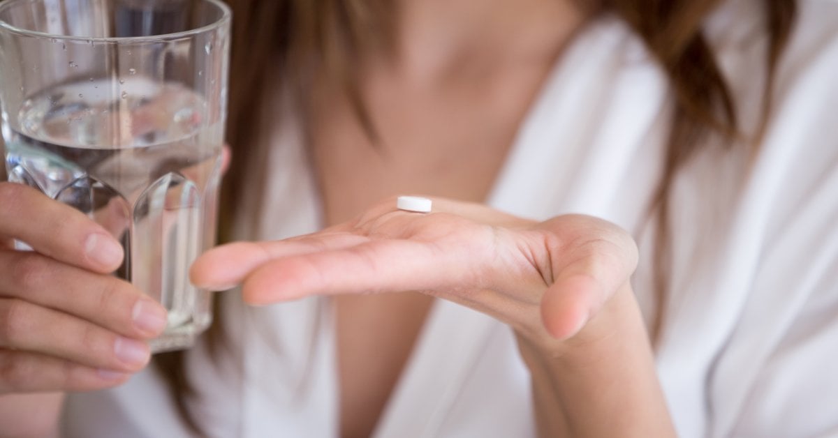 A person holds a glass of water in their right hand. They hold a white pill in the palm of their left hand.