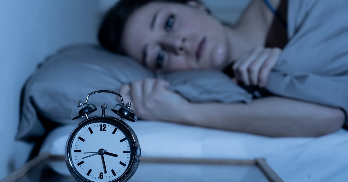 A woman lies awake in bed at night with an alarm clock nearby, showing signs of anxiety and insomnia.
