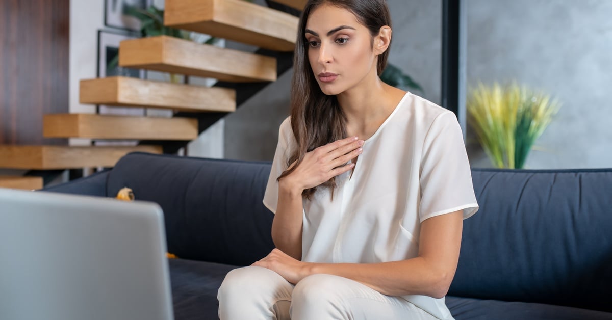 A woman sits on a couch with her hand on her chest, appearing to manage anxiety during an online therapy session.
