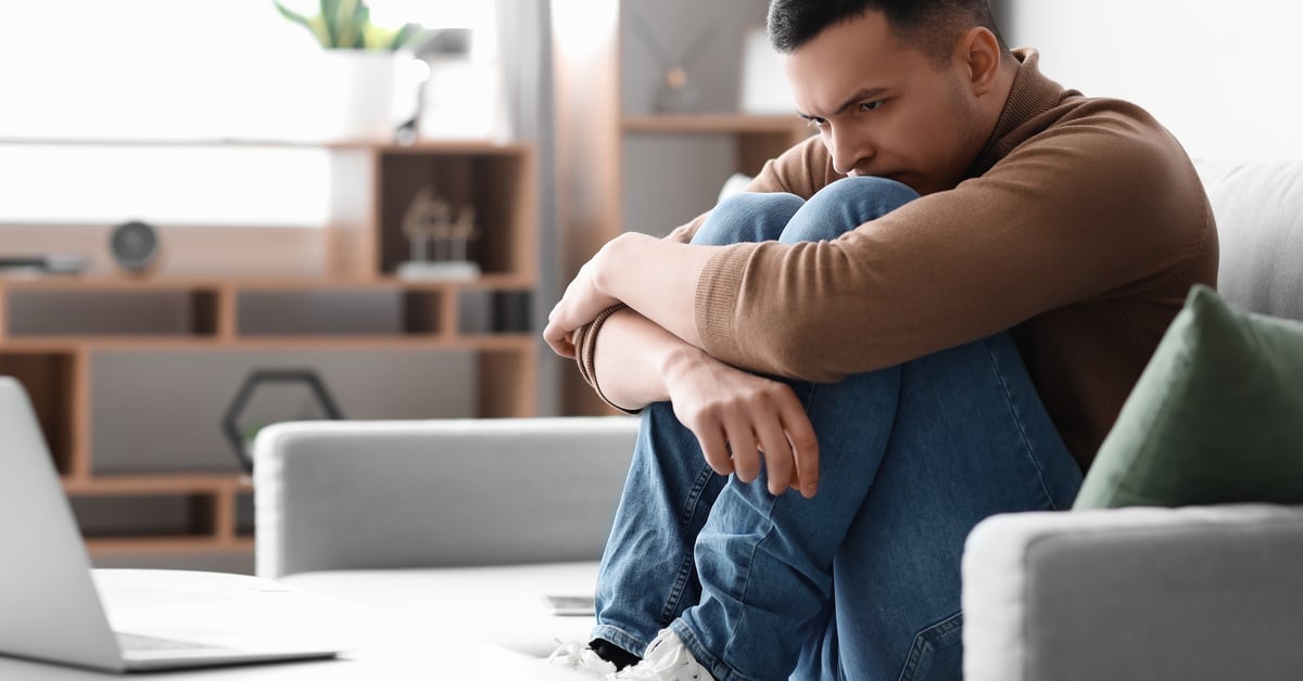 A man sits on a couch hugging his knees with a worried expression, showing signs of anxiety while at home.