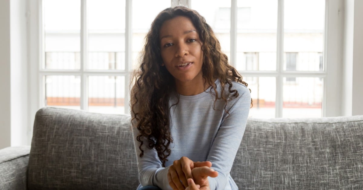 A young Black woman gestures while speaking during an online talk therapy session from her living room.
