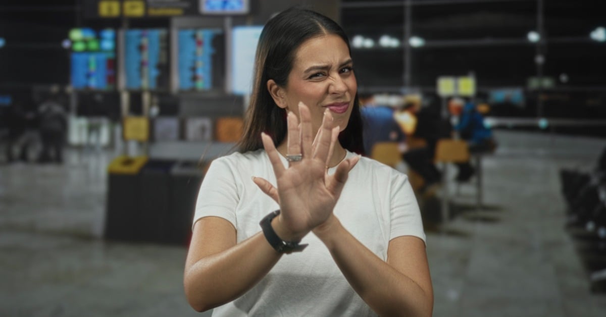 A woman raises both hands in a stop gesture, showing a disgusted expression while setting boundaries in an airport terminal.
