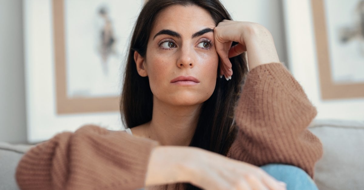 A woman sits on a couch with her knees drawn to her chest. She rests her head on one hand and looks to the side.