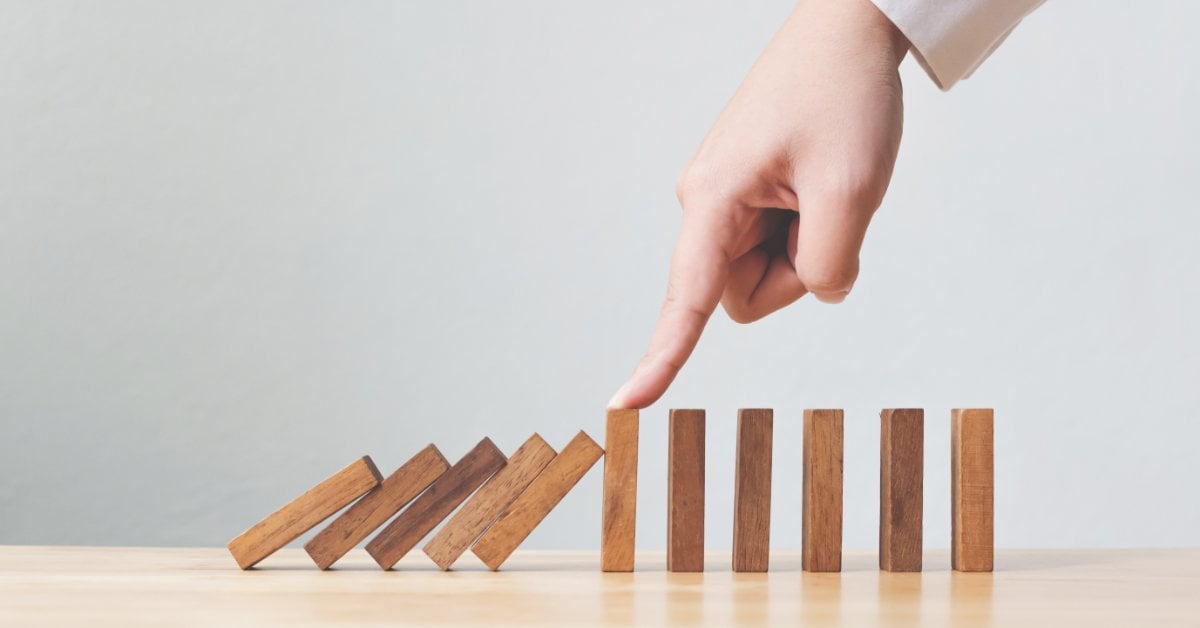 A hand presses down on one wooden block in the middle of a row of them to stop it from knocking down the rest.