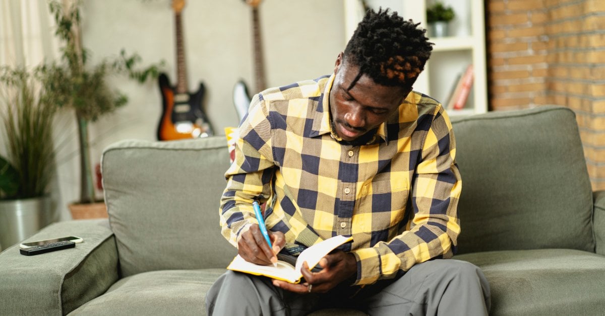 A man wearing a yellow plaid shirt sits on a couch and writes in a notebook. Two guitars hang on the wall behind him.