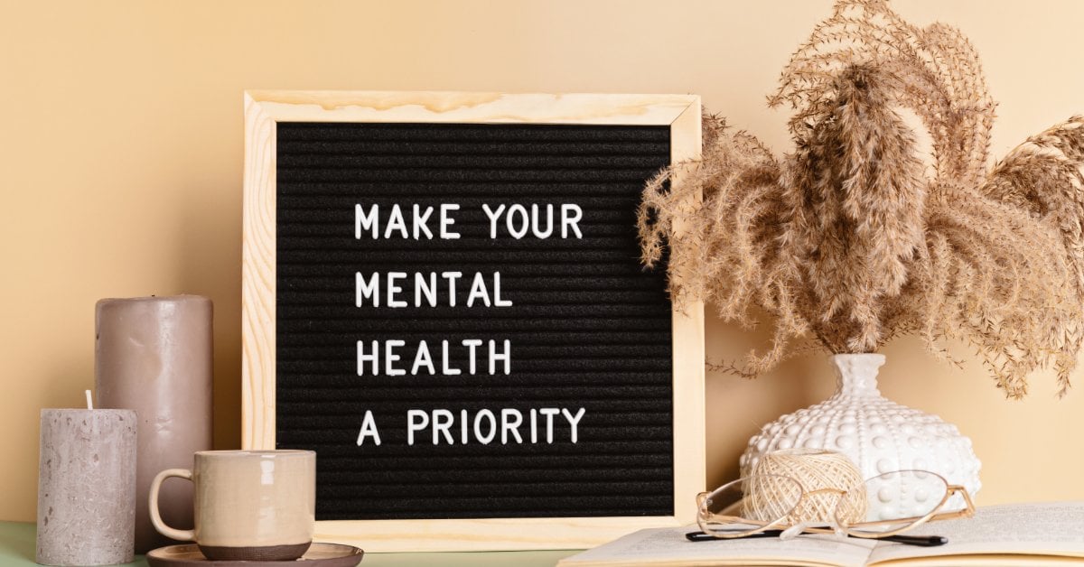A black letter board on a table reads “Make your mental health a priority” beside candles, a mug, and dried plants.