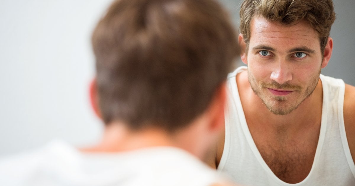 A man in a white tank top looks at himself in a mirror. The back of his head and shoulders are in the foreground.