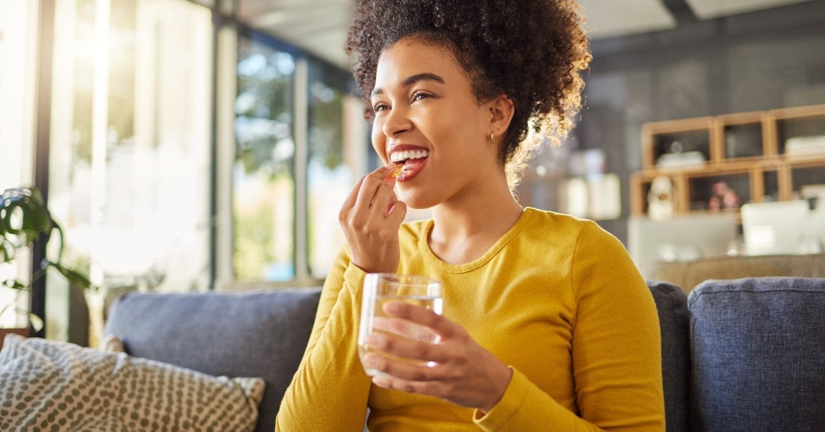 A smiling woman sitting on a couch holds a glass of water. With her other hand, she brings a pill to her open mouth.