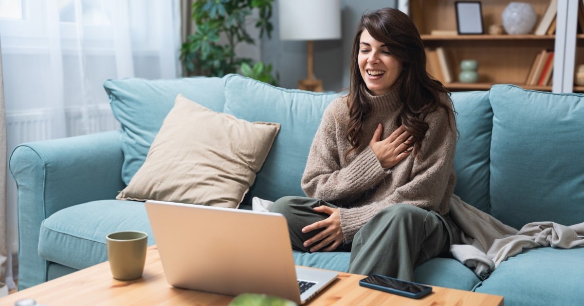 A woman sits on a blue couch in front of an open laptop. A mug and a smartphone are on the coffee table in front of her.