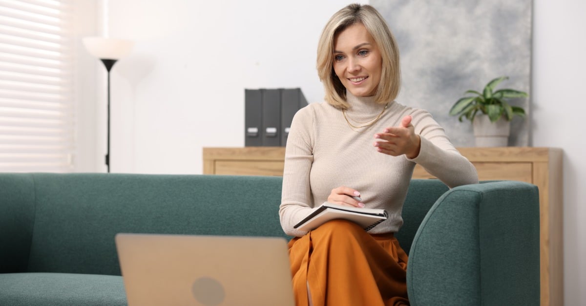 A woman sits with a notebook in her lap and gestures toward a laptop. A cabinet and file folders are behind her.