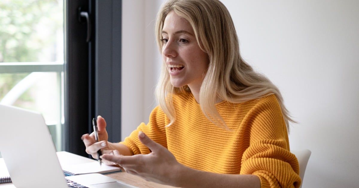 A woman gestures as she talks and looks at a laptop screen. She sits at a table with a notebook near a large window.