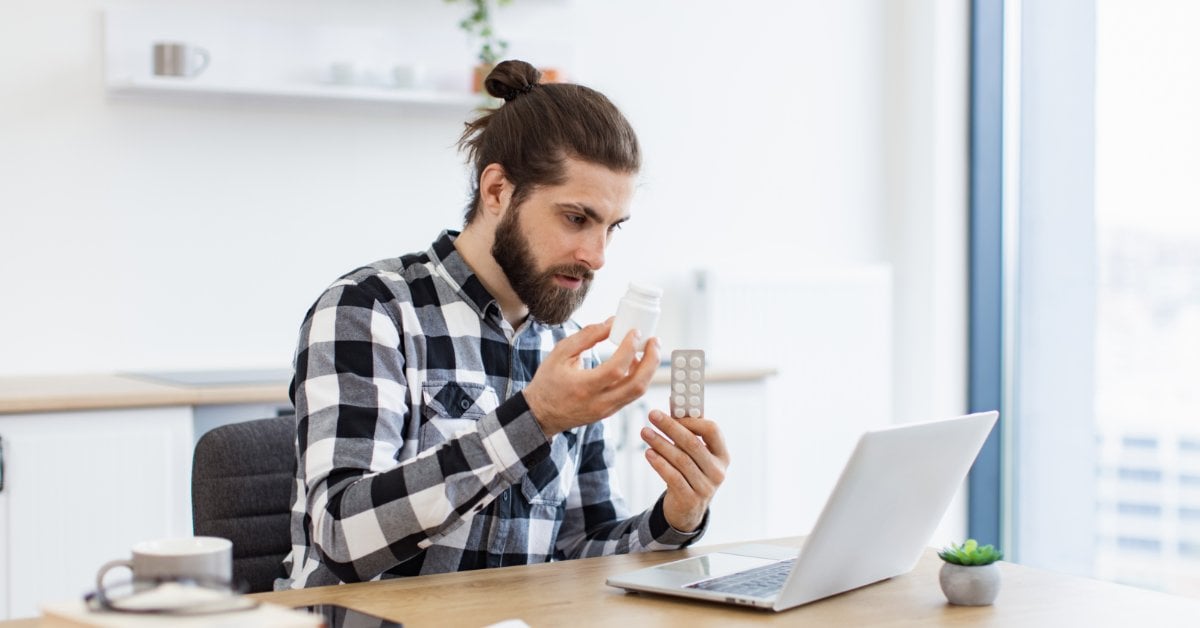 A man sits in front of an open laptop and holds up a medication bottle in one hand and a blister pack in the other.