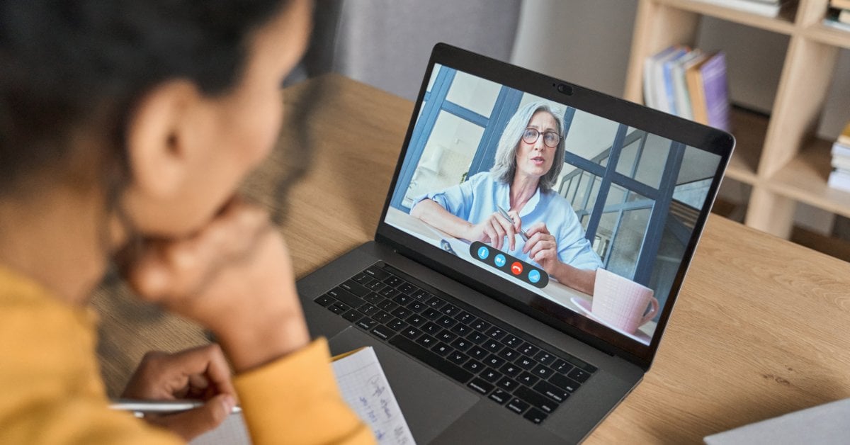 A person in a yellow sweater takes notes while looking at a laptop during a video call. A woman on the screen talks.
