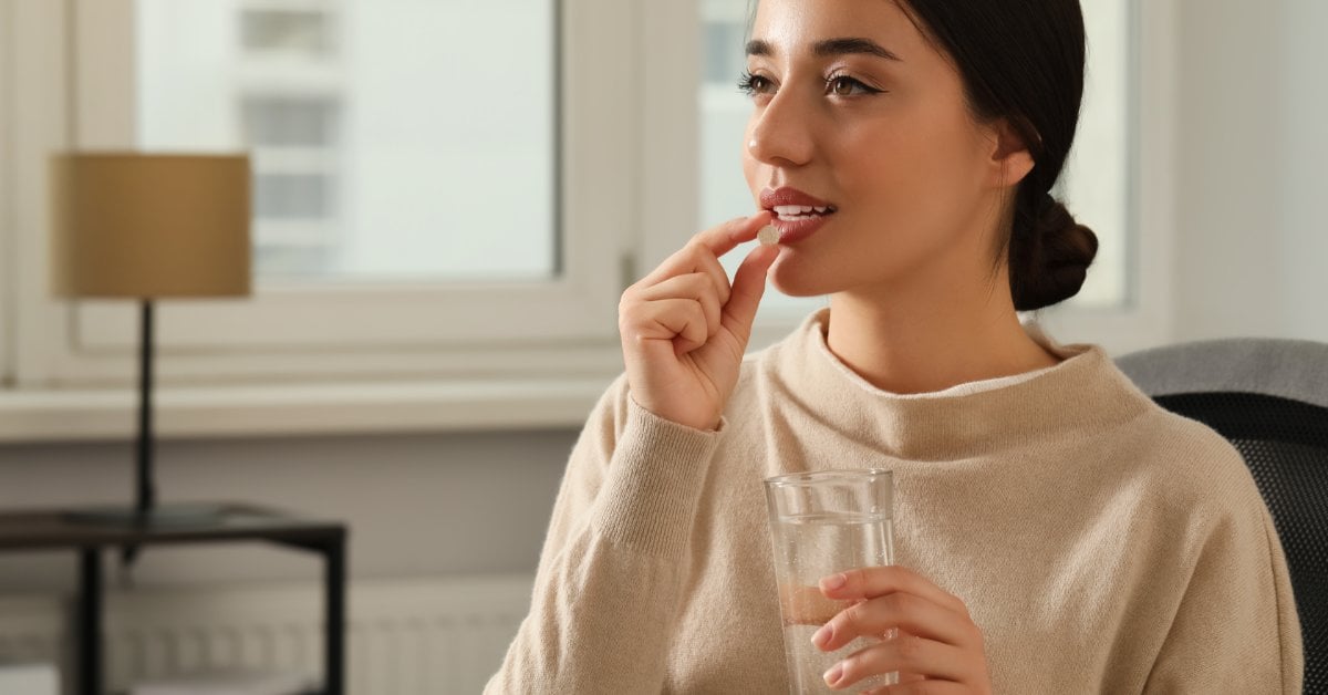 A woman holds a glass of water in one hand while bringing a pill to her mouth. She sits indoors near large windows.