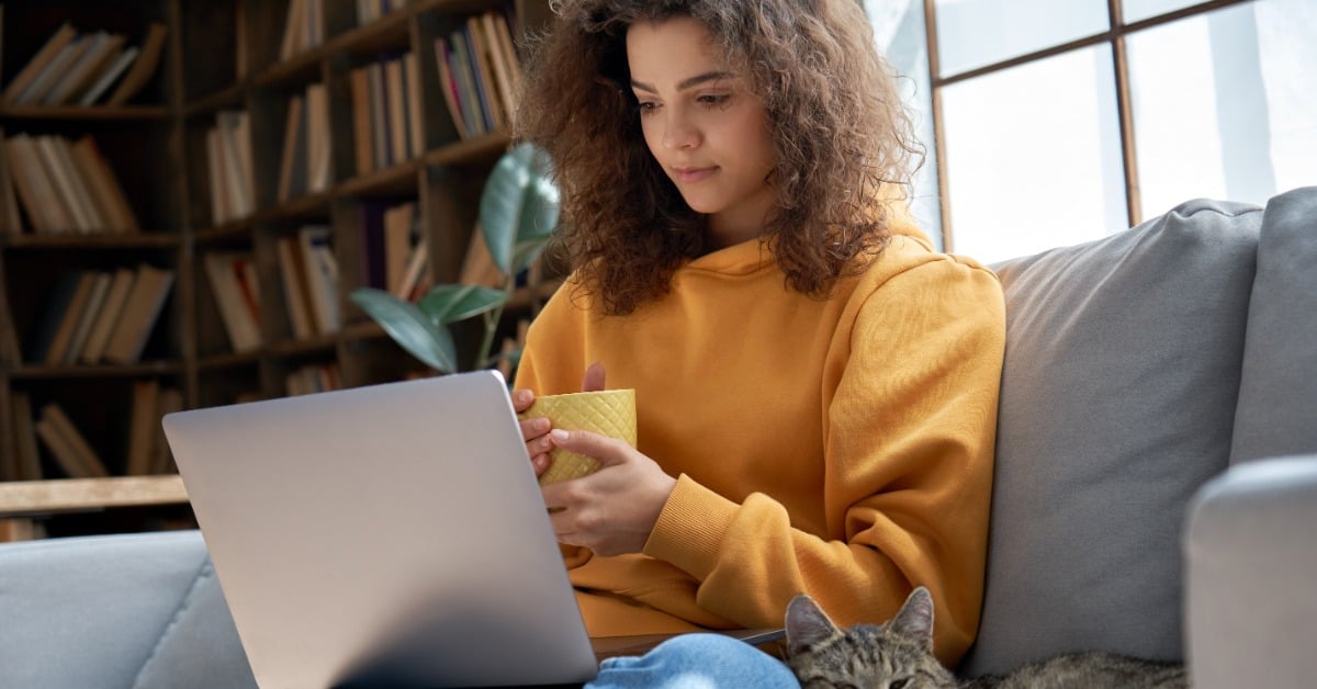 A woman sits on a couch with a laptop on her lap and a mug in her hands. A cat lies beside her on the cushion.