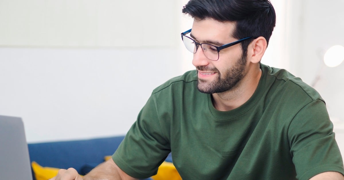 A man wearing glasses sits with a laptop and an open notebook. He holds a pencil and looks at the laptop screen.