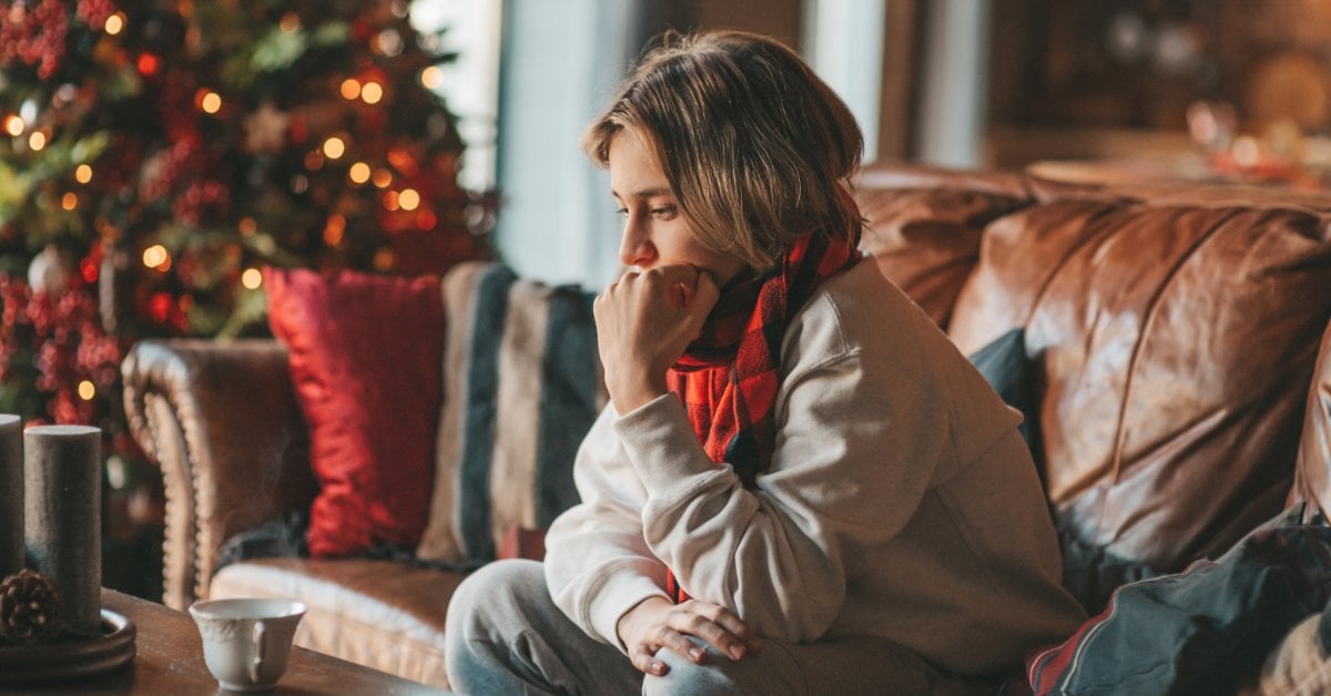 A woman leans forward on a couch, her chin against her fist. A Christmas tree with glowing lights stands behind her.