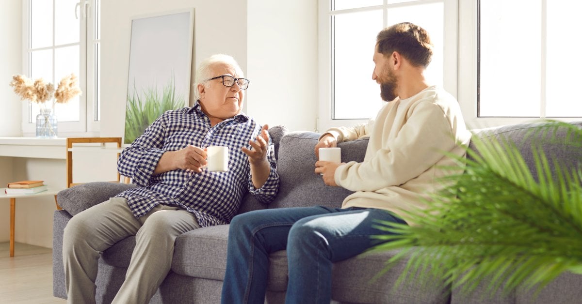 An older man wearing glasses gestures while talking to a younger man on a gray couch. Both men hold white mugs.