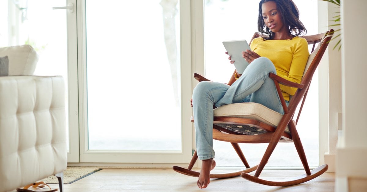A woman sits in a wooden rocking chair while using a digital tablet. Sunlight shines from the glass doors behind her.
