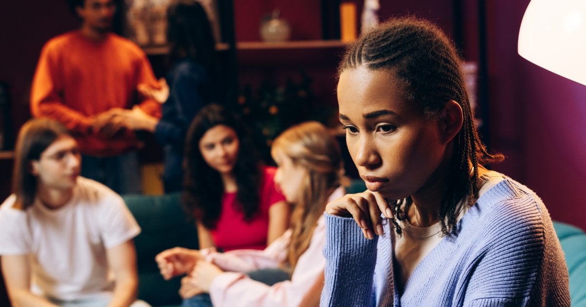 A woman rests her chin on her hand, looking down and away from people sitting and talking on a couch behind her.