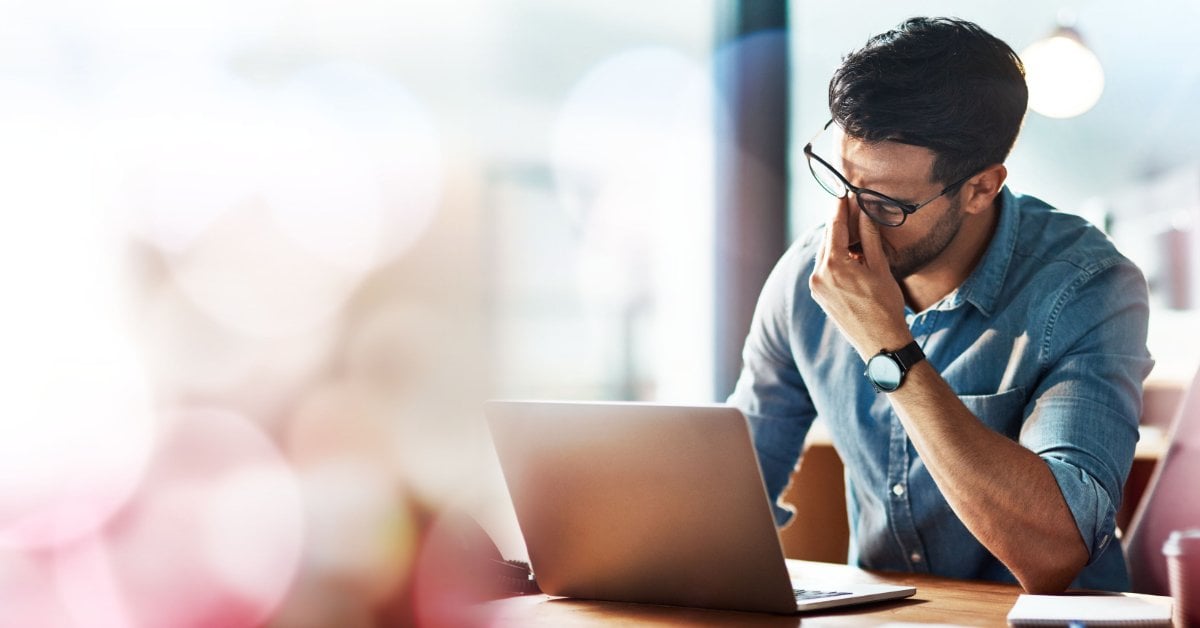 A man with a laptop squeezes the bridge of his nose under his glasses. Blurred bokeh circles are in the foreground.