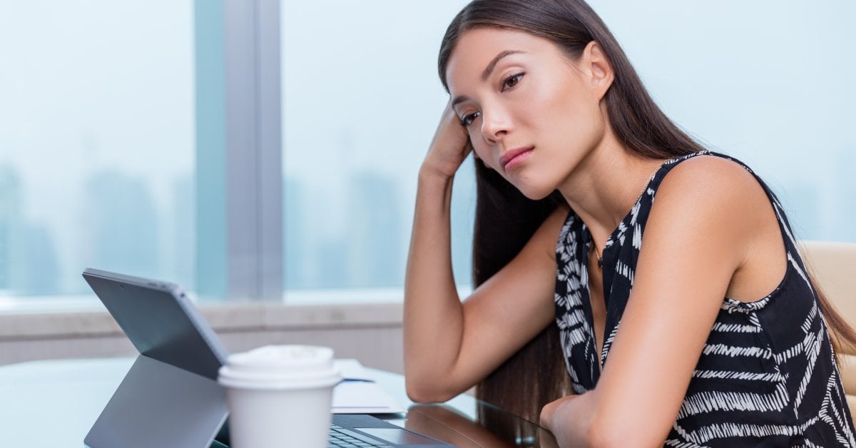 A woman rests her head on one hand while looking off to the side. A tablet and a disposable cup are in front of her.